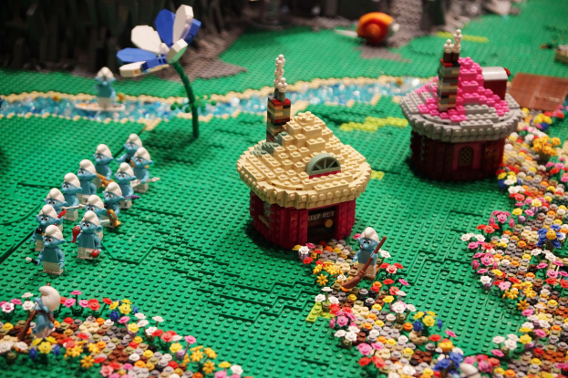 Ein LEGO-Modell einer Kleinstadt mit bunten Blumen, Spielzeugen, Häusern, Pflanzen und anderen Gegenständen, vor einem leicht unscharfen Hintergrund.