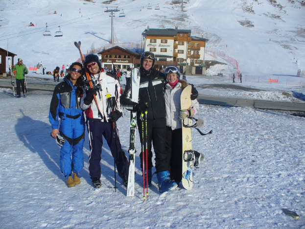 Vier Personen posieren für ein Foto mit Skating Pads und Werkzeugen, mit Gebäuden und anderen Menschen im Hintergrund, vor einer schneebedeckten Berglandschaft.