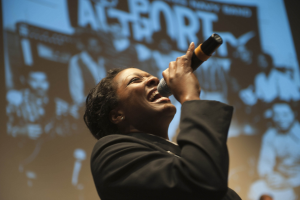 Eine Frau in einem schwarzen Kleid mit einem Dutt singt leidenschaftlich in ein Mikrofon auf der Bühne, ihr Gesicht von Scheinwerfern beleuchtet und ihre Augen vor emotionaler Ausdruckskraft geschlossen.