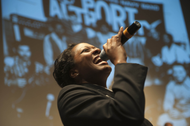 Eine Frau in einem schwarzen Kleid mit einem Dutt singt leidenschaftlich in ein Mikrofon auf der Bühne, ihr Gesicht von Scheinwerfern beleuchtet und ihre Augen vor emotionaler Ausdruckskraft geschlossen.