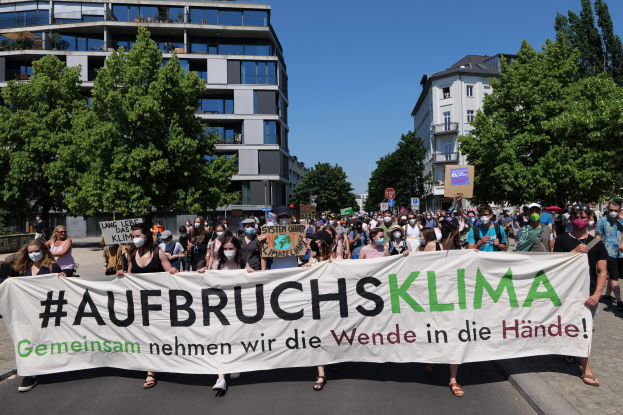 Eine Gruppe von Menschen in Masken hält ein Banner mit der Aufschrift "Aufbruchsklima" vor einem Gebäude mit Bäumen und einem klaren Himmel.