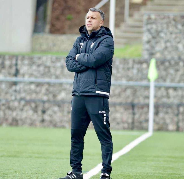 Ein Mann steht auf einem Fussballfeld mit verschränkten Armen, trägt eine schwarze Jacke und Hose und hat im Hintergrund einen Zaun, Treppe und Wand.