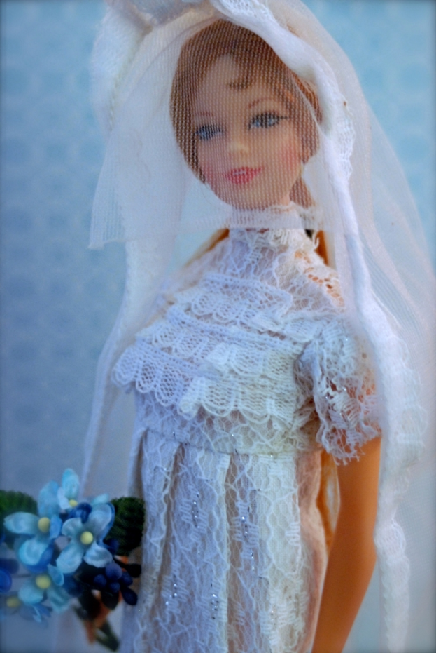 Eine Puppe in einem Kleid, Schleier und mit einem Strauß in der Hand, vor einem Hintergrund.
