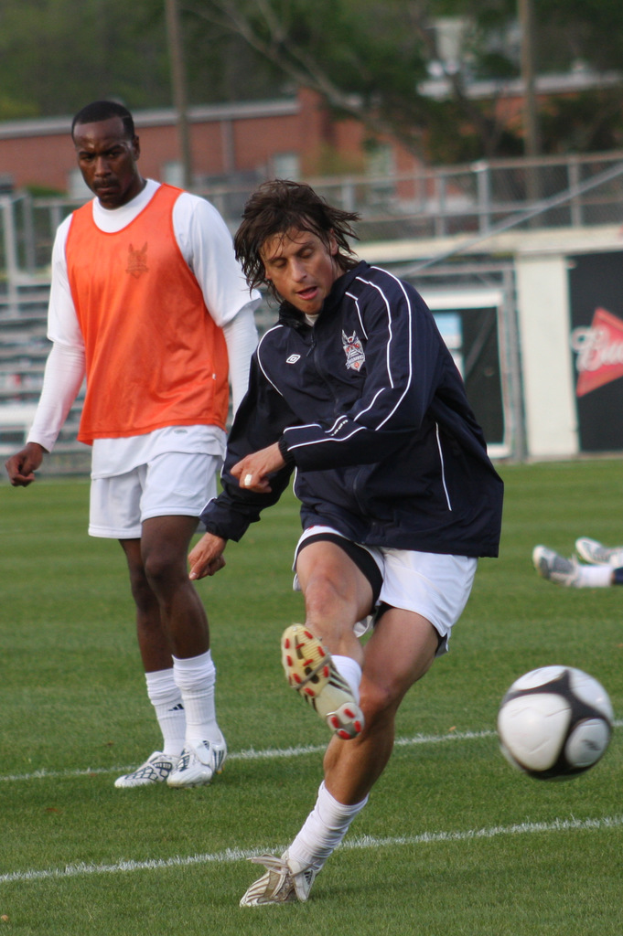 Ein Fußballspieler in einer schwarzen Jacke und weißen Shorts schießt den Ball, während ein anderer Spieler in einer orangefarbenen Jacke hinter ihm steht und ein Zaun mit Bäumen im Hintergrund zu sehen ist.