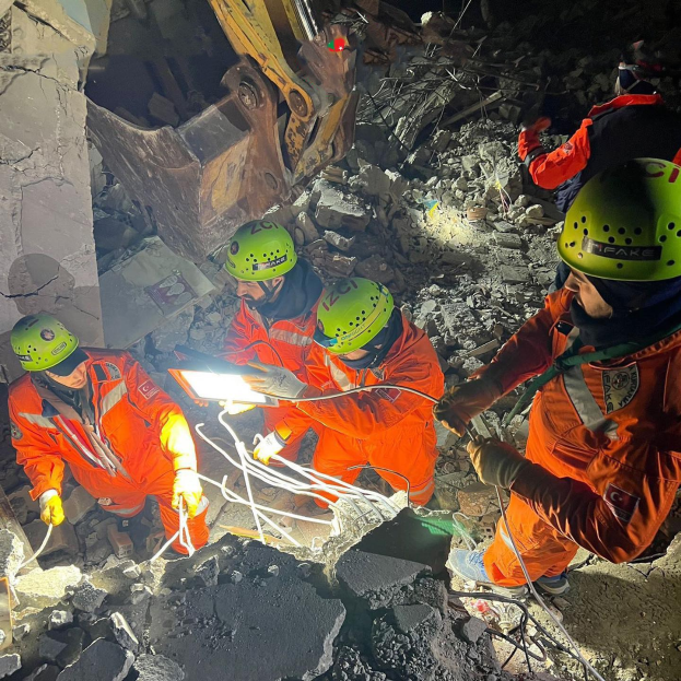 Eine Gruppe von Rettungskräften in orangefarbenen Uniformen und grünen Helmen beseitigt Trümmer von einem eingestürzten Gebäude, mit Werkzeugen, mit einem Kran im Hintergrund.