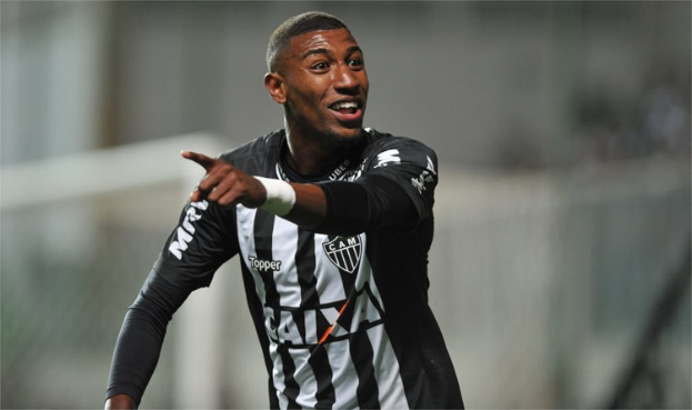 Ein Fußballer in einem schwarzen und weißen T-Shirt lächelt und feiert nach einem Tor, mit einem unscharfen Hintergrund.