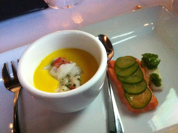 Eine Schüssel Suppe mit einem Löffel und einer Gabel daneben und ein Servierteller mit Fleisch, das mit Gurkenscheiben garniert ist.